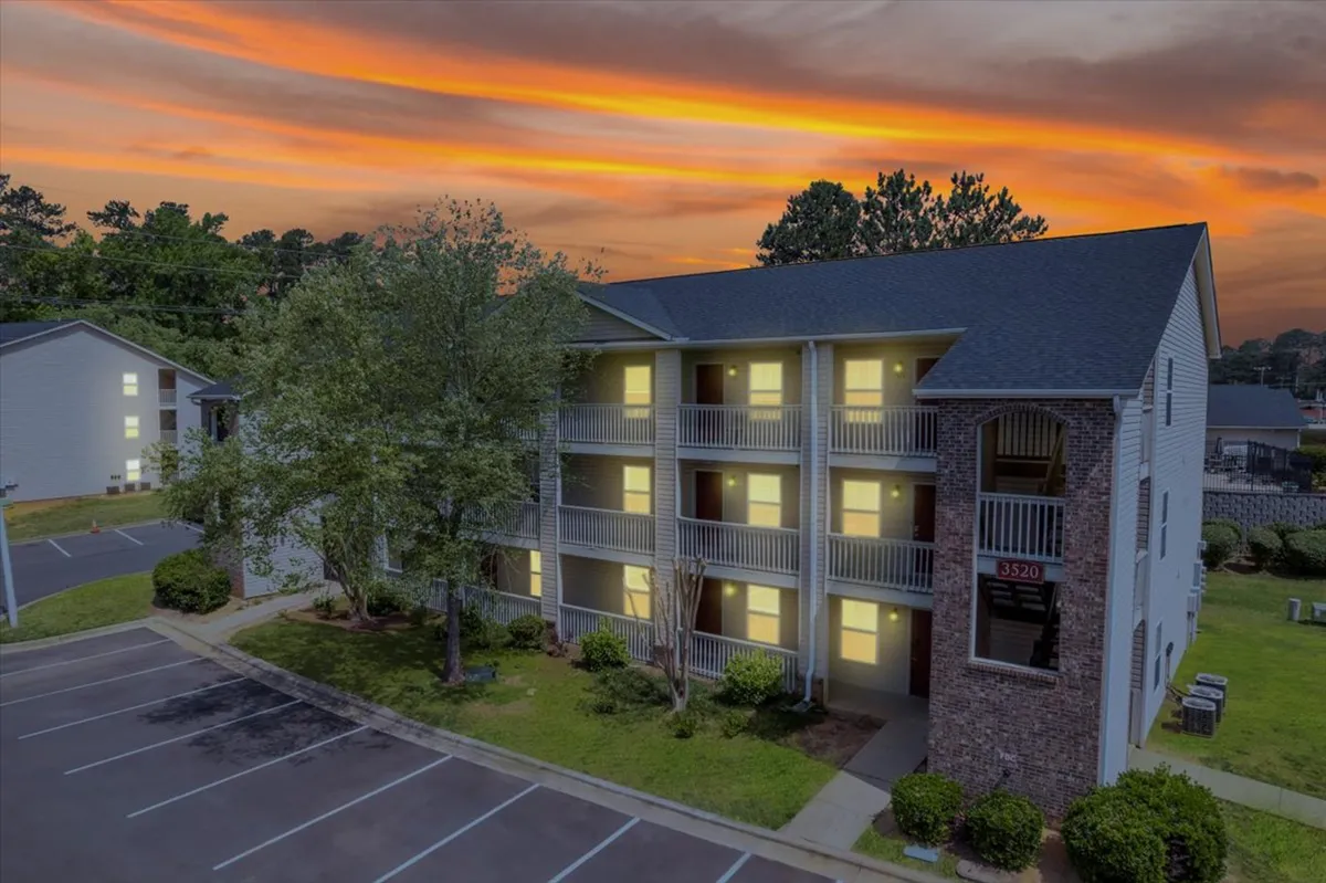 This exterior shot captures the three-story apartment building during a vibrant sunset, with lush greenery surrounding the property and inviting balconies visible. The calm evening atmosphere is enhanced by the warm, welcoming glow of natural light.