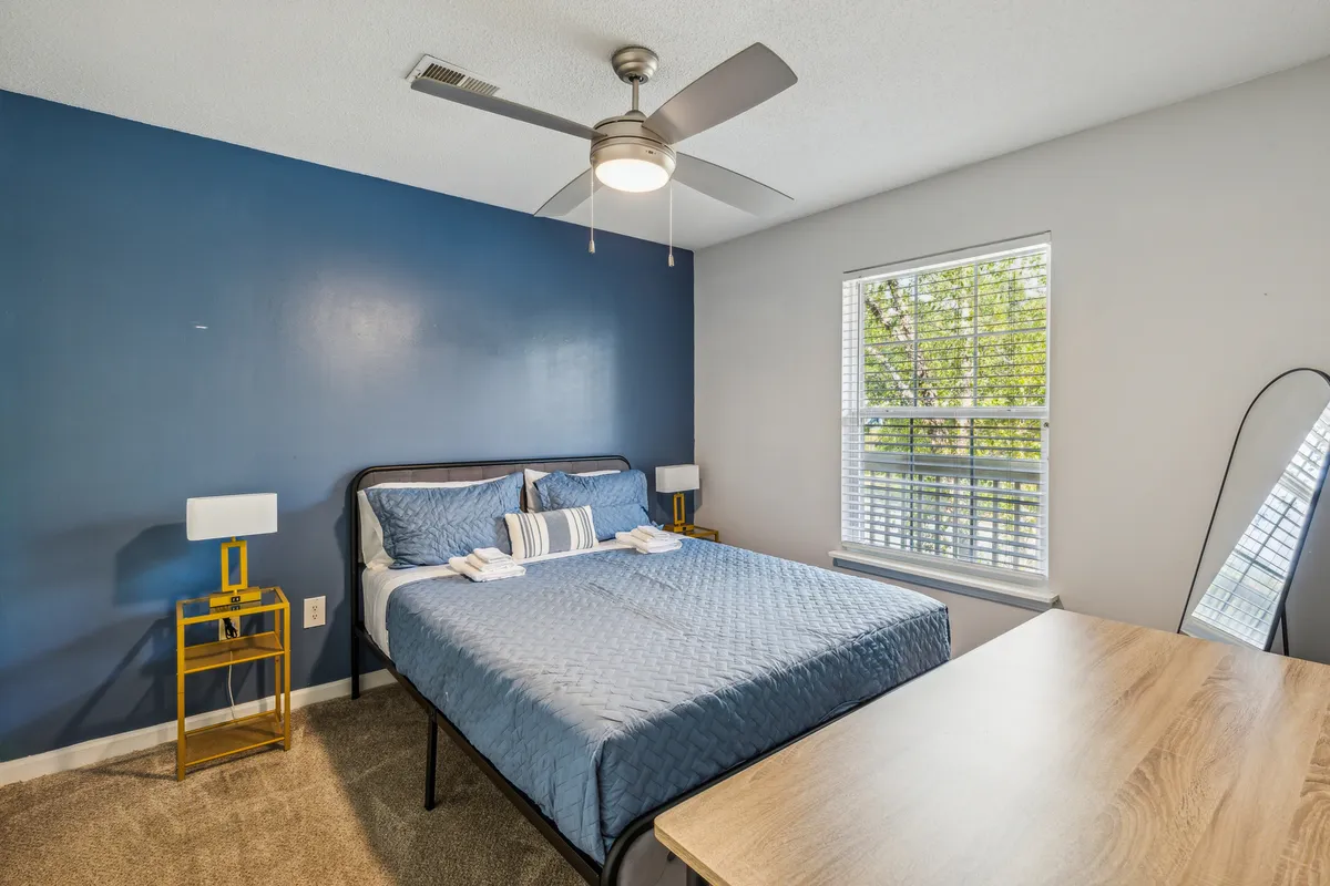 A serene bedroom featuring a plush queen bed with inviting blue bedding, a gold side table, ample natural light pouring in through a large window, and a wooden desk tucked to the side. The space is styled with a calming palette, offering both comfort and functionality.