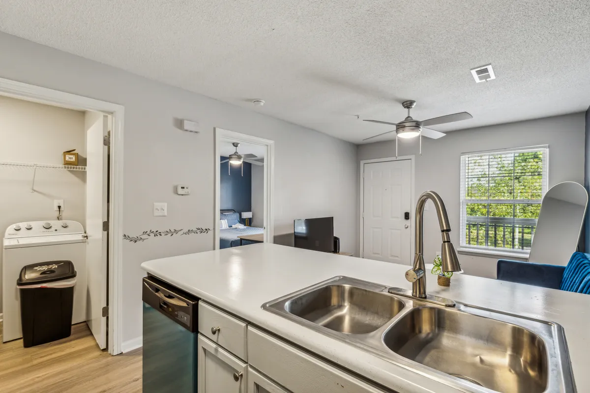 A cozy kitchen space with a central island, a visible washing machine tucked neatly to the side, and a window letting in natural light. The scene conveys a lived-in, homey atmosphere where doing laundry is seamlessly integrated into daily life.