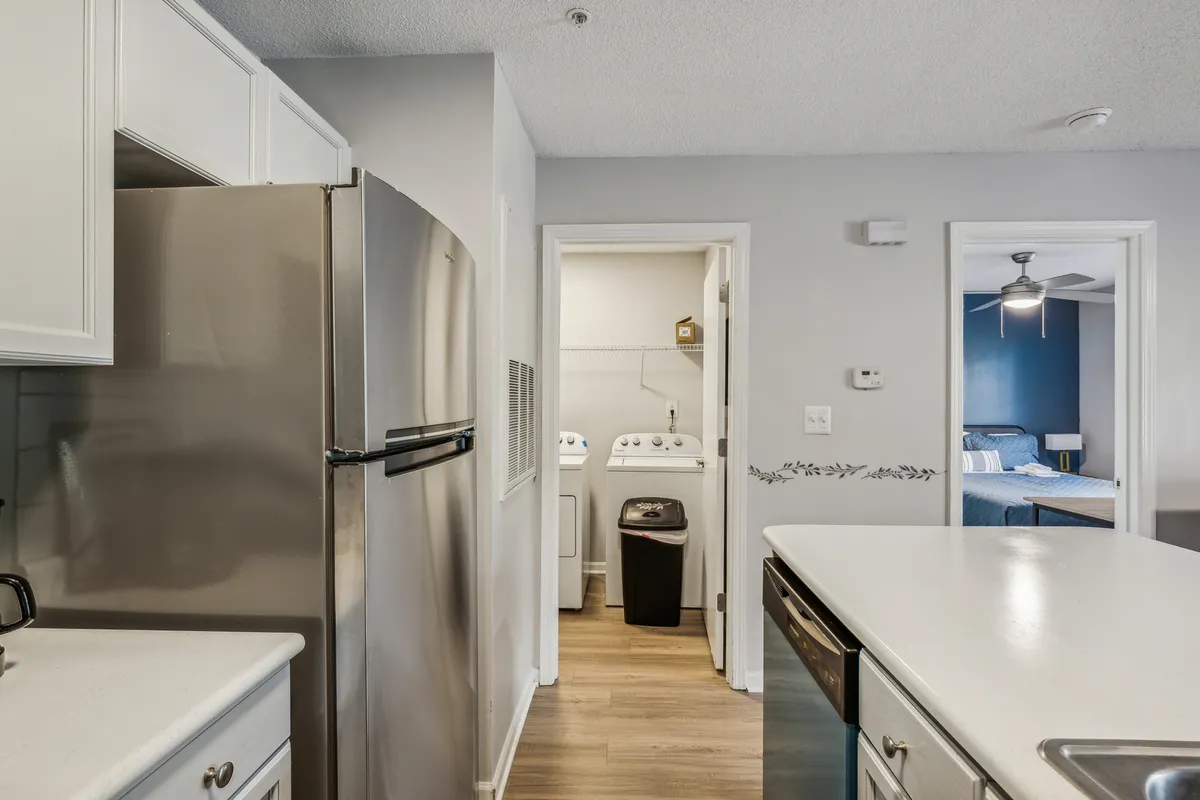 A modern kitchen featuring a stainless steel refrigerator, white countertops, and a clear view into the adjacent laundry area. The bright, functional design emphasizes ease of access and contemporary living.