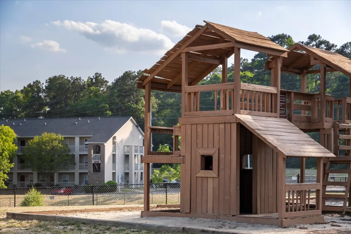 An inviting exterior view of the apartment building, showcased behind a lively playground surrounded by lush greenery. The scene captures a residential environment that feels both safe and welcoming, with spaces for community interaction and relaxation outside.