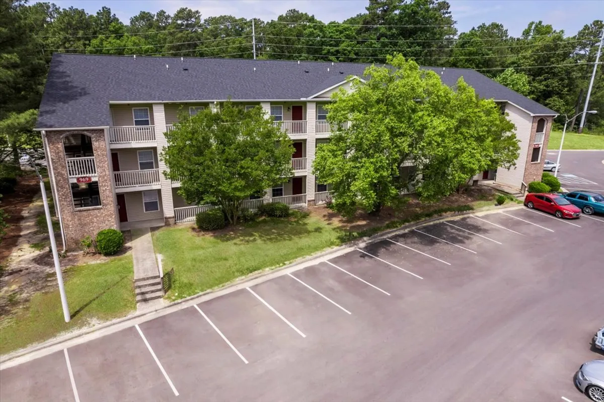 This exterior image showcases a welcoming brick apartment building with expansive balconies shaded by a mature tree in front. Residents can be seen relaxing on the balconies, and there is convenient parking right outside. The scene suggests easy access and a pleasant, tranquil environment just steps from home.