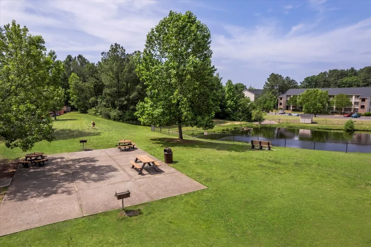 An inviting picnic area beside a serene pond, surrounded by mature trees and benches. The space is designed for outdoor relaxation and gatherings, with a sense of calm and natural beauty.