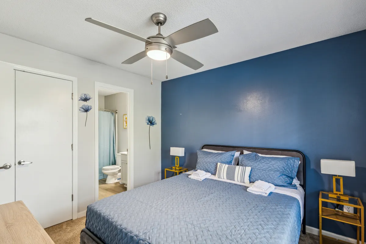 The image showcases a cozy bedroom with a plush bed dressed in inviting blue bedding, set against a bold blue accent wall. Bright yellow nightstands add a cheerful pop of color, while a ceiling fan promises comfort. Natural light fills the room, creating a bright, airy, and restful atmosphere.