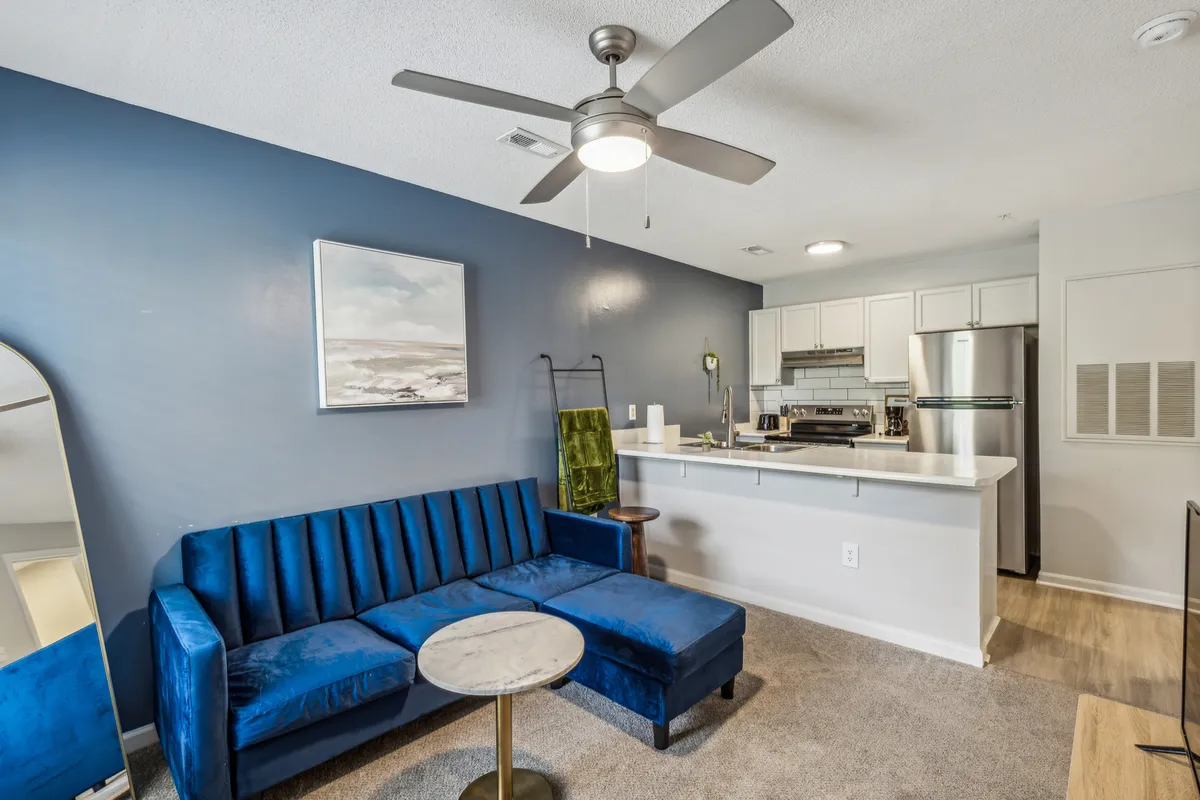 A view of a blue velvet couch with a round coffee table, modern kitchen area in the background, and abstract wall art, bathed in natural light. The open concept design promotes easy flow from entry to lounge.