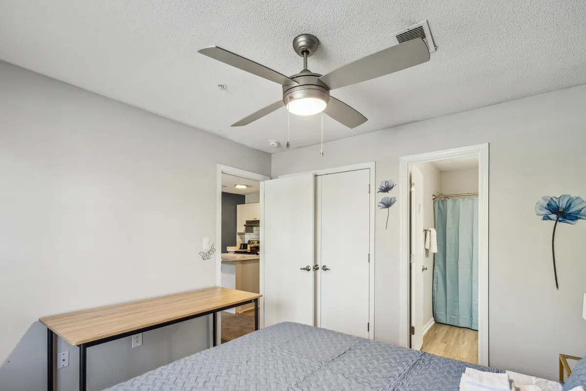 A cozy bedroom scene with a queen bed, a ceiling fan overhead, a desk for work or reading, closet doors for storage, and delicate blue flower wall decor that enhances the calming ambiance. The room feels comfortable and private, ideal for winding down after a busy day.